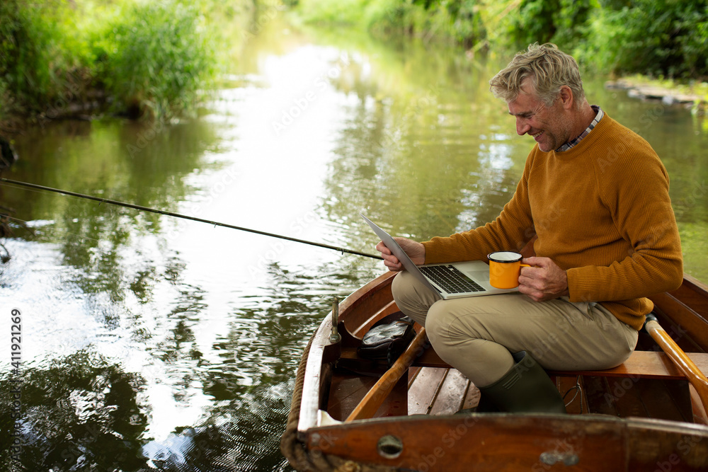Man fly fishing and using laptop with coffee in boat on river Man fly fishing and using laptop with coffee in boat on river