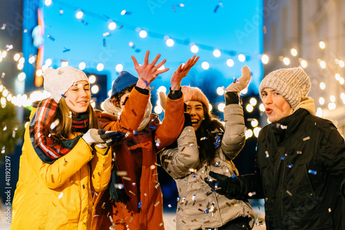 Happy attractive young men women wearing woolen hats having fun and jumping with colorful confetti outdoors. Festive, chill, celebration, holiday concept.