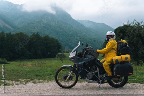 A girl in a yellow raincoat  shoe covers and a white helmet. Motorcyclism and travel. Sightseeing tour. Top of the Mountains. A gray day with thunderclouds. Copy space  biker s outfit.