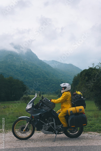 A girl in a yellow raincoat, shoe covers and a white helmet. Motorcyclism and travel. Vertical photo. Top of the Mountains. A gray day with thunderclouds. Difficult test and biker's outfit.