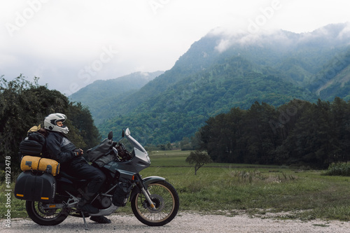 A motorcycle driver with motorbike take a rest, Adventure vacation, biker dressed in raincoat. sealed bag, water resistant, overalls. Mountains road trip, side bags equipment. copy space