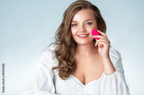 Woman applying foundation using cosmetic sponge, beauty blender. Photo of woman with perfect makeup on light blue background. Beauty and Skin care concept. Plus size model.