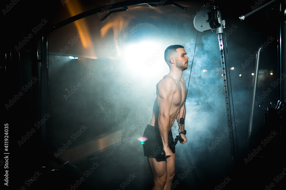 Fototapeta premium Muscular man working out in gym doing exercises with dumbbells at triceps, strong male naked torso abs, athlete doing triceps exercise arm muscles atmospheric smoke in the gym