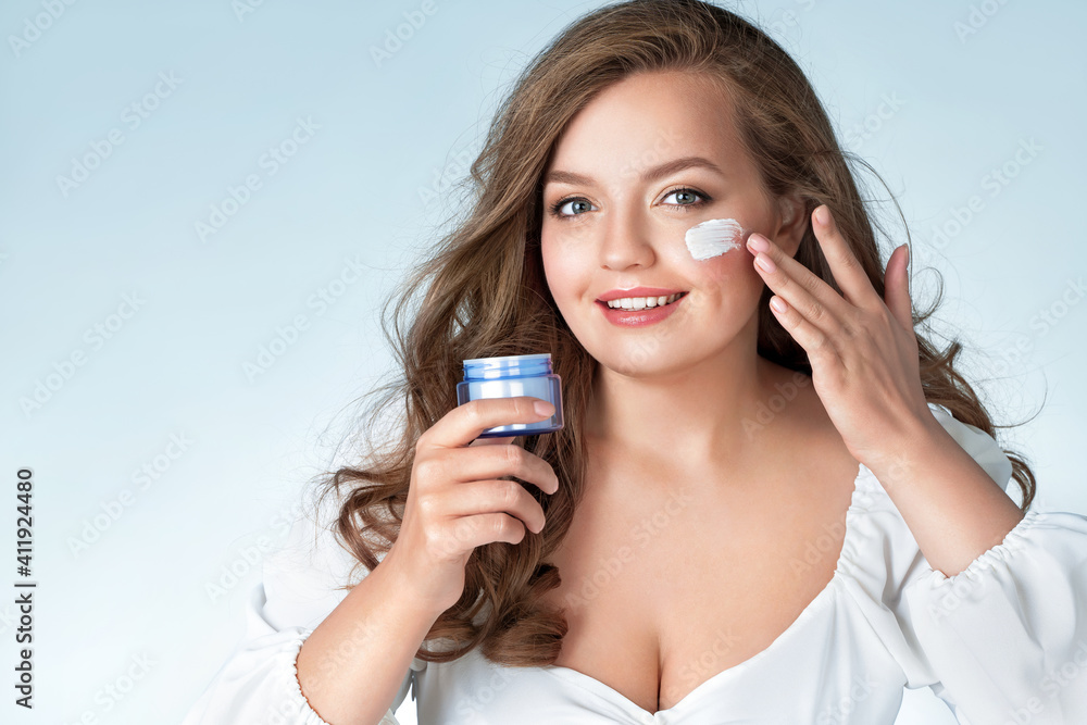 Beautiful woman applying moisturizer cream on her face. Photo of woman with perfect skin on light blue background. Beauty and Skin care concept. Plus size model. Beautiful woman applying moisturizer cream on her face. Photo of woman with perfect skin on light blue background. Beauty and Skin care concept. Plus size model.