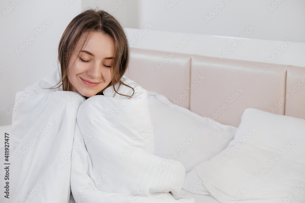 Pretty young smiling Caucasian woman waking up in the morning and stretching in her bed and looking content. Covered in white linen. Pretty young smiling Caucasian woman waking up in the morning and stretching in her bed and looking content. Covered in white linen.