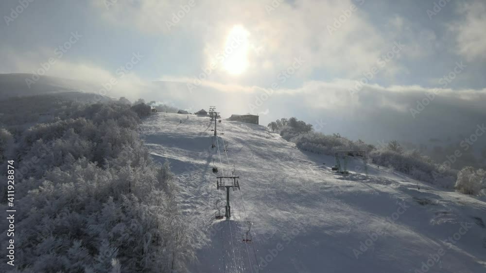 Flight over the winter ski lift. People in ski suits ride a ski lift up a snowy mountain. Aerial view of ski sift for transportation skiers on snowy ski slope. Flight over the winter ski lift. People in ski suits ride a ski lift up a snowy mountain. Aerial view of ski sift for transportation skiers on snowy ski slope.