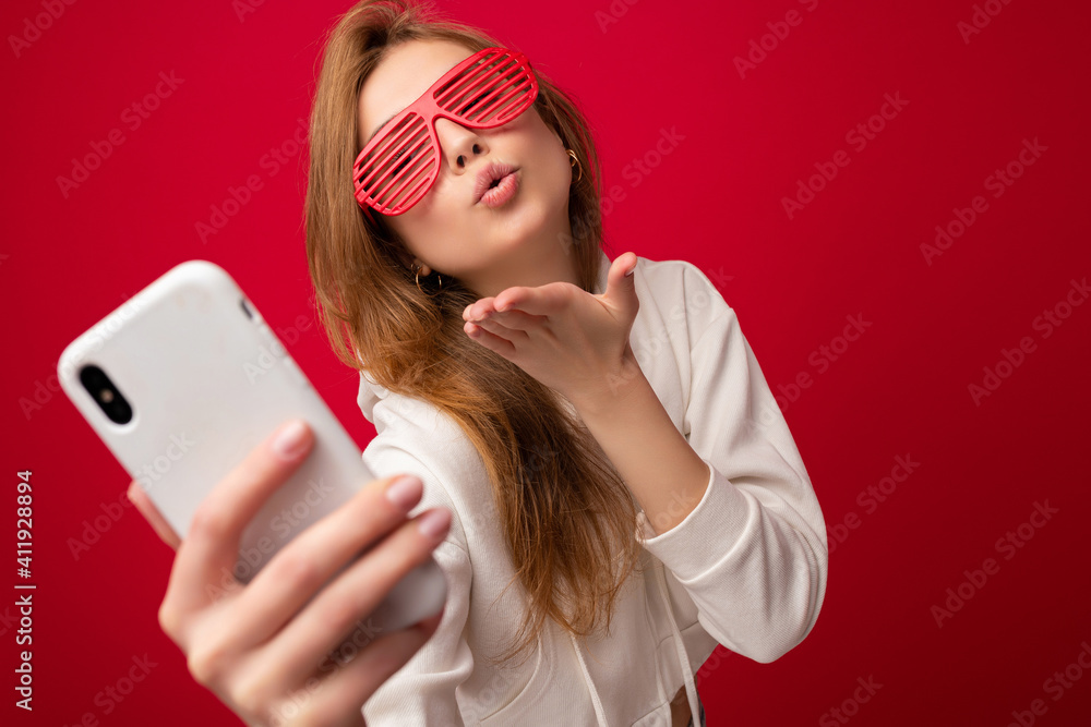 Naklejka premium Closeup portrait of attractive positive smiling young blonde woman wearing stylish white hoodie and funny colorful glasses standing isolated over colourful background holding and using mobile phone