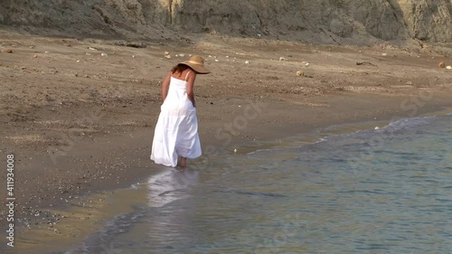 A woman alone in a white dress on a deserted beach walks on a mud path and dry land next to the sea 