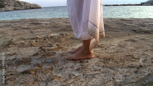 A woman alone in a white dress on a deserted beach walks on a mud path and dry land next to the sea 