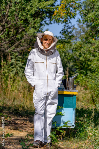 Beekeeper poses to the camera in protective uniform on a small apiary farm. Apiculture concept. Summer day at apiary.