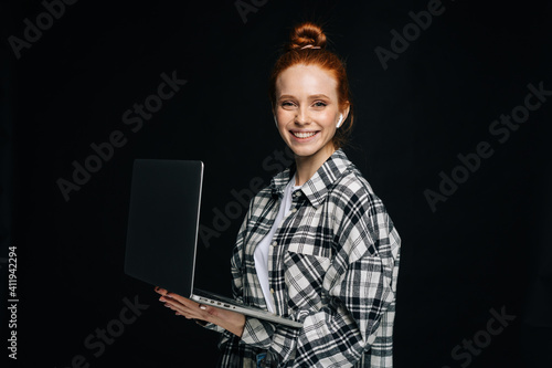 Happy charming young business woman or student holding laptop computer and looking at camera on isolated black background. Pretty redhead lady model emotionally showing facial expressions, copy space.