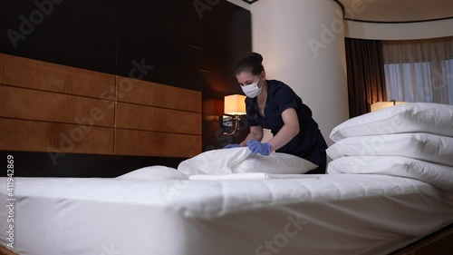 Woman hotel employee in uniform, face mask and gloves preparing room for new visitors during covid infection spread. Chambermaid putting on clean pillowcases while working in hotel bedroom