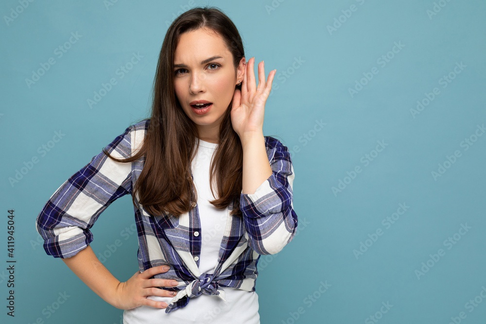 Photo of young concentrated attractive pretty brunet woman with sincere emotions wearing stylish check shirt standing isolated on blue background with empty space and listening to something by putting Photo of young concentrated attractive pretty brunet woman with sincere emotions wearing stylish check shirt standing isolated on blue background with empty space and listening to something by putting