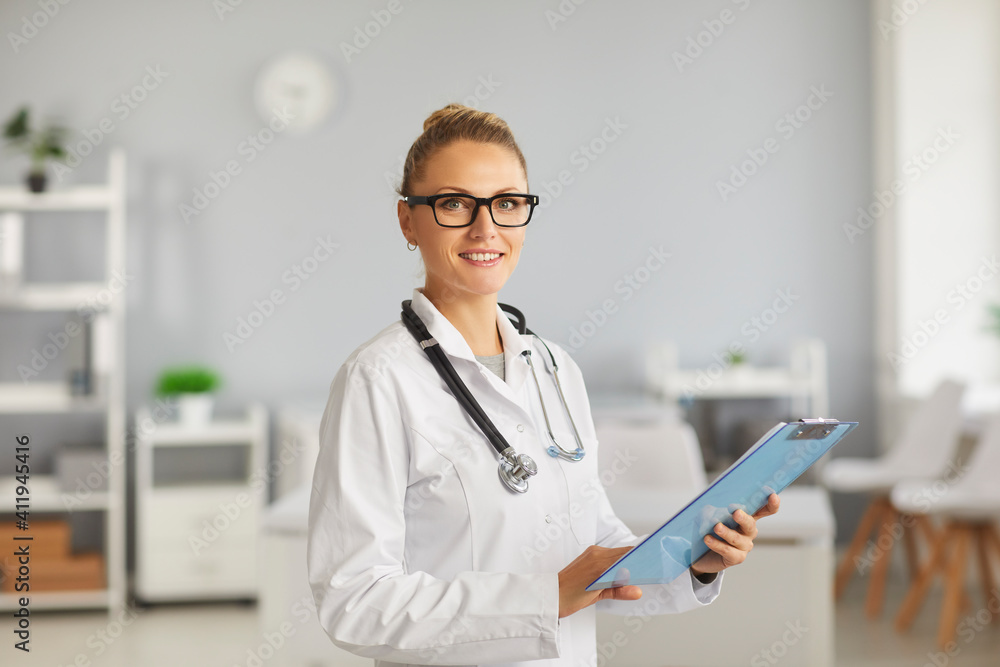 Profile staff. Portrait of smiling female doctor with glasses, therapist, general practitioner looking at camera, standing in hospital with clipboard in hands. Concept of professional doctors. Profile staff. Portrait of smiling female doctor with glasses, therapist, general practitioner looking at camera, standing in hospital with clipboard in hands. Concept of professional doctors.