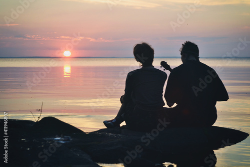 Valokuva Guy and  girl in love are sitting on the shore of the lake at sunset and sing a song under the ukulele
