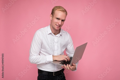 Side profile photo shot of handsome confident blonde man holding computer laptop typing on keyboard wearing white shirt looking at camera isolated over pink background
