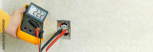 Male electrician checks the voltage of an outlet on a white wall with a multimeter in the house close up. Banner photo