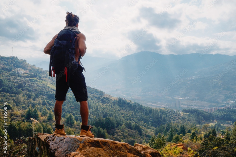 Obraz premium Young man standing with long hair without shirt and big backpack on rock in the mountain looking the city