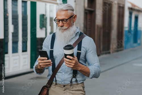 an older man in hipster clothes and glasses and a long white beard drinks coffee while walking down the street and looks at his mobile phone focus on head