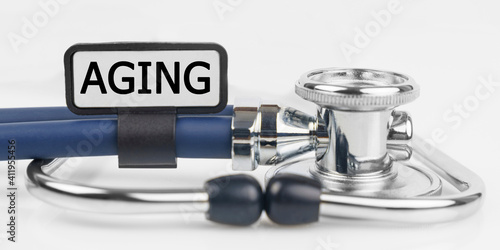On the white surface lies a stethoscope with a plate with the inscription - AGING