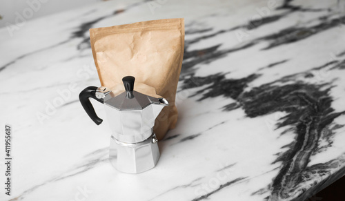 A silver coffee maker Moka and a craft bag of roasted coffee beans on a marble table