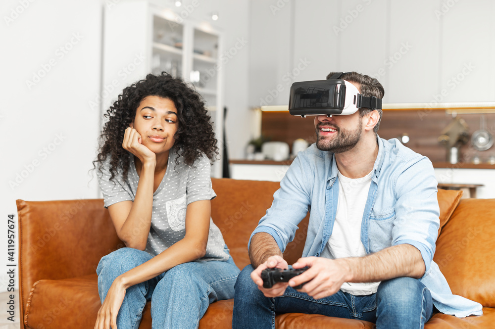 Interracial couple sitting on the couch, guy playing video game without  paying attention to young African American girlfriend, she is annoyed by  her addicted boyfriend in vr headset and with joystick Stock