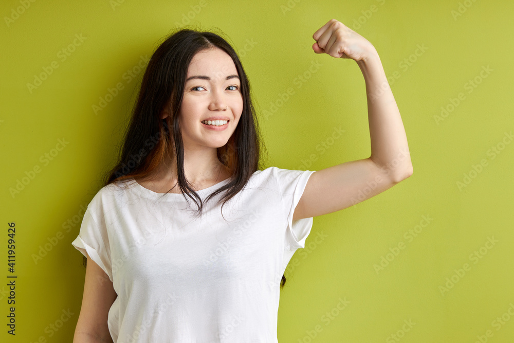 young woman raises arm and shows biceps has happy expression demonstrates her achievements after training in gym dressed casually isolated on green background. Look at my muscles young woman raises arm and shows biceps has happy expression demonstrates her achievements after training in gym dressed casually isolated on green background. Look at my muscles