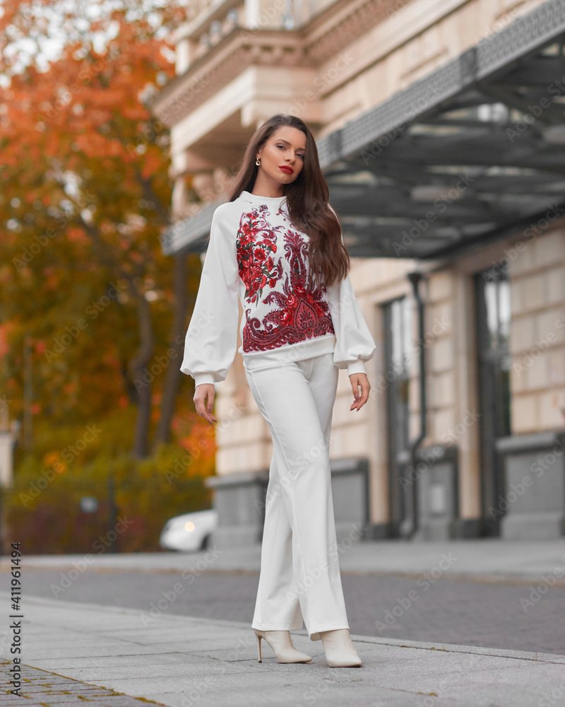 Elegant caicasian woman with long straight brunette hair in white sporty glamour costume and high heels walking city street on autumn day
