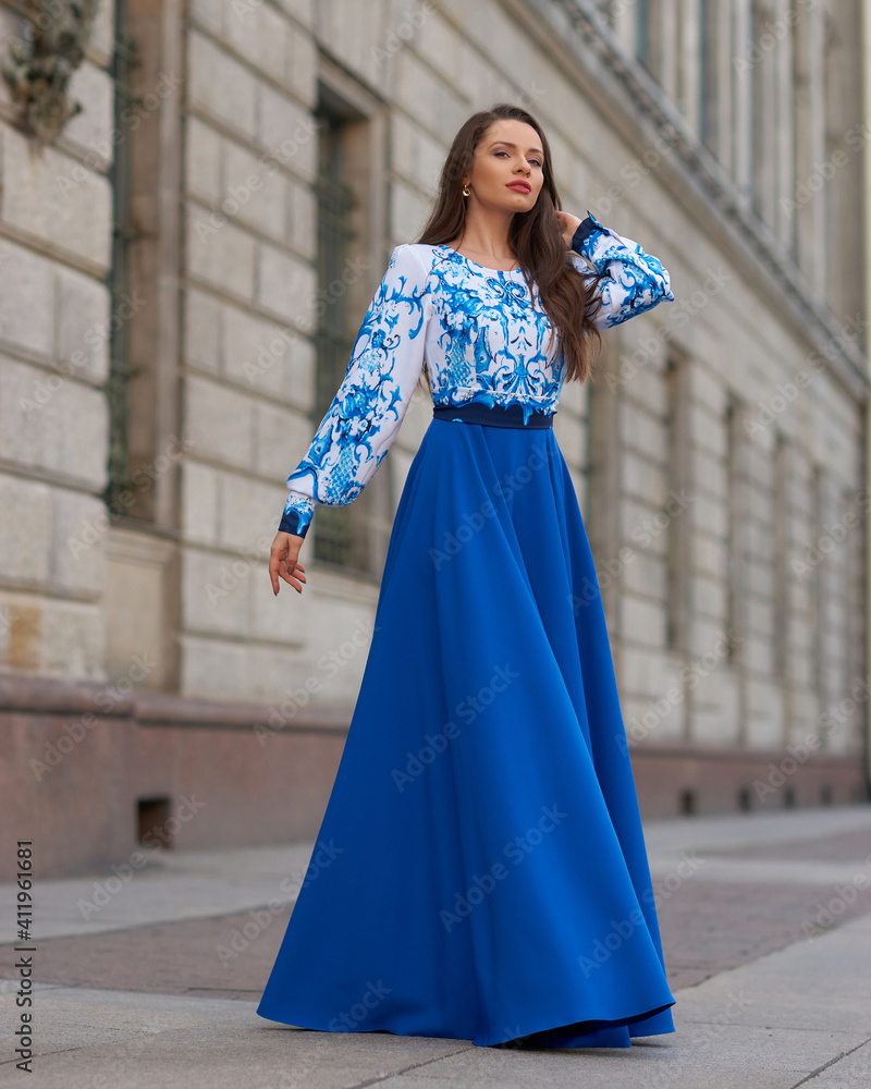 Fototapeta premium Elegant caucasian woman with long straight brunette hair in blue and white stylish colorful dress walking city street on a bright day