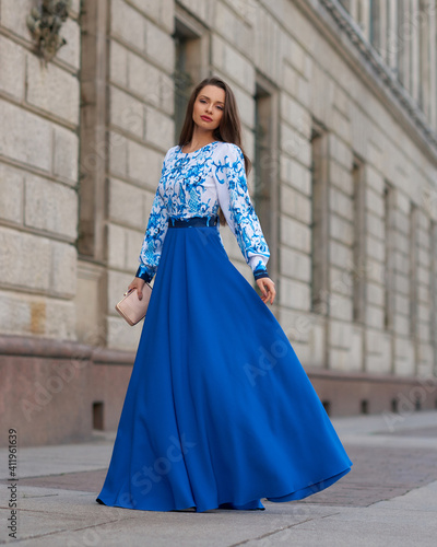 Elegant caucasian woman with long straight brunette hair in blue and white stylish colorful dress walking city street on a bright day