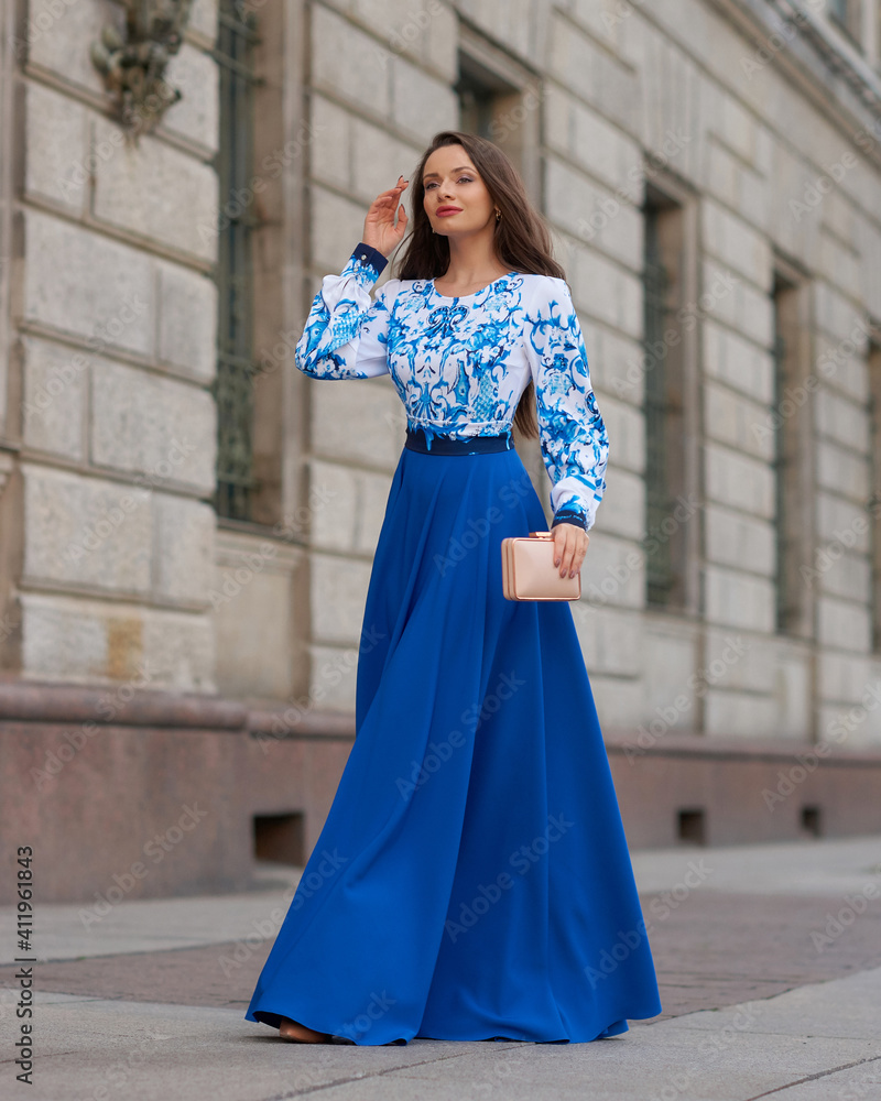 Elegant caucasian woman with long straight brunette hair in blue and white stylish colorful dress walking city street on a bright day