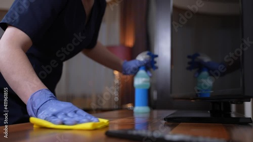 Close-up of gloved housekeeper's hands spraying sanitizer and carefully wiping surface of table and TV-set while cleaning house. Masked maid in uniform and gloves using sprayer and duster during work