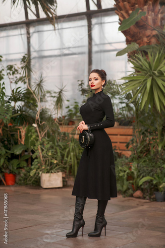 Glamorous young woman in black midi dress posing in greenhouse