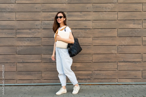 Full length photo shot of young charming beautifyl brunet woman going in the street near brown wall in white t-shirt and light blue jeans white sneakers with black female bag holding computer laptop photo