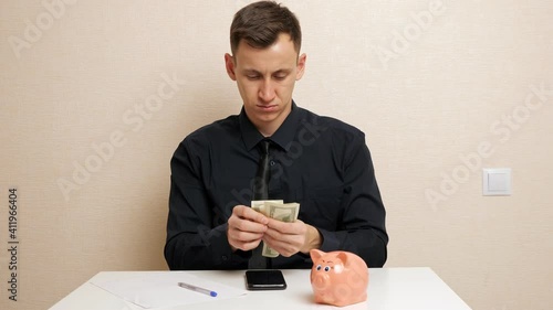Man takes dollar banknotes out of shirt breast-pocket and writes family expenses on white paper sitting near piggy bank at beige wall closeup photo