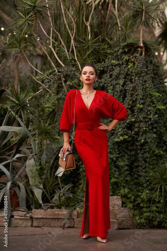 Stunning woman in red evening maxi dress posing in greenhouse