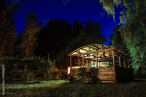 Wooden gazebo on a green field in the forest against the background of the night blue sky © ISliM