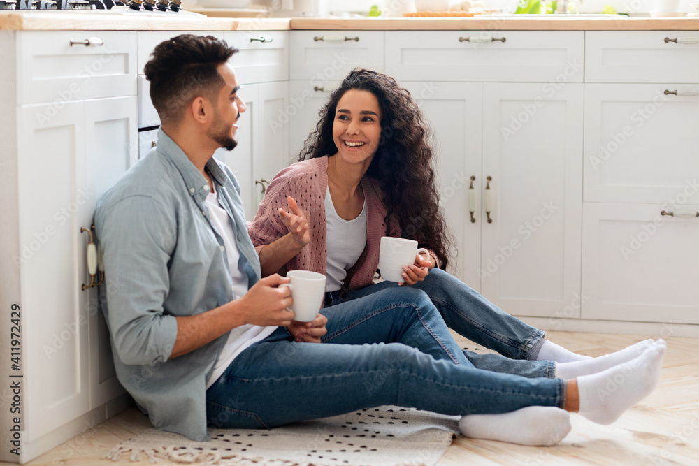 Cheerful Arab Couple Drinking Coffee In Kitchen, Sitting With Cups On Floor Cheerful Arab Couple Drinking Coffee In Kitchen, Sitting With Cups On Floor