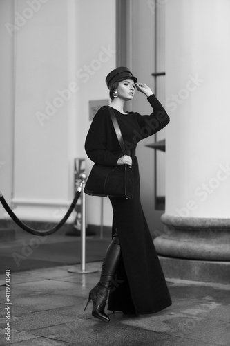 Elegant woman in stylish black maxi dress and cap posing outside exquisite building