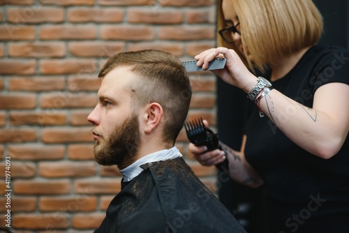 Professional hairdresser woman doing styling of handsome guy with electric hair clipper at barber shop