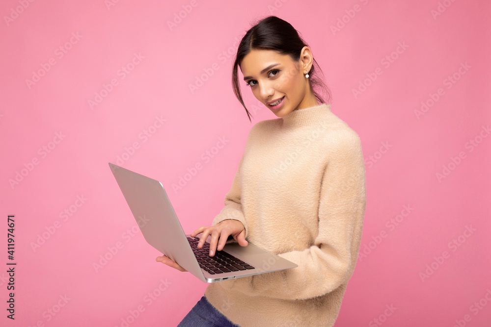 Naklejka premium Photo of Beautiful young brunette woman wearing beige sweater holding netbook computer typing text on keyboard looking at camera isolated over pink wall background