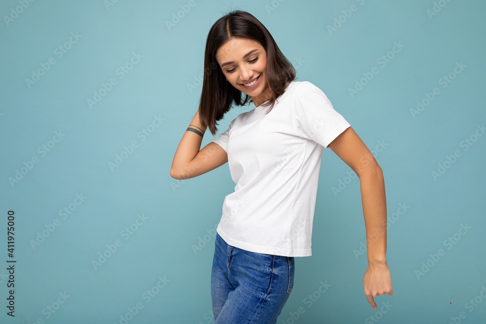 Photo of positive smiling joyful young beautiful brunette woman with sincere emotions wearing casual white t-shirt for mockup isolated over blue background with empty space and dancing Photo of positive smiling joyful young beautiful brunette woman with sincere emotions wearing casual white t-shirt for mockup isolated over blue background with empty space and dancing