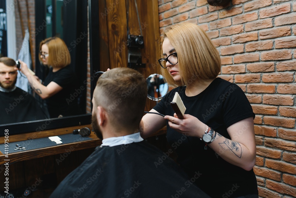 Serving client in Barbershop. Professional barber girl, female hairdresser making modern haircut for a man sitting in barber shop chair. Focus on a girl. Hairdressing, shaving, trimming, grooming
