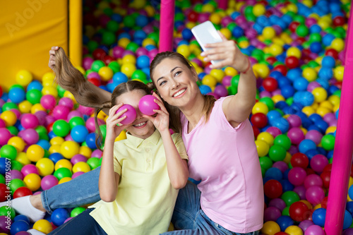 Beautiful young woman with her teen daughter taking selfie together at ball pond in kids amusement centre