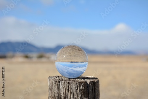 Glass lens ball showing inverted reflection of mountains and farm fields with blue sky and clouds through a crystal ball on a fence post.