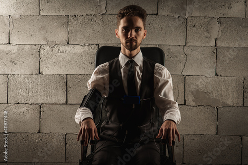 Concentrated, pensive, man in a gray room sitting on a chair, testing on a polygraph, has sensors for passing a polygraph, sitting on a gray brick wall background. The concept of truth, polygraph photo