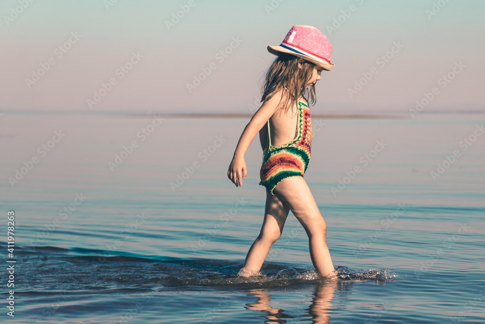 Naklejka premium A cute girl in a knitted swimsuit and a pink hat walks in shallow water along the seashore. Copy space. Toned minimalism photo.