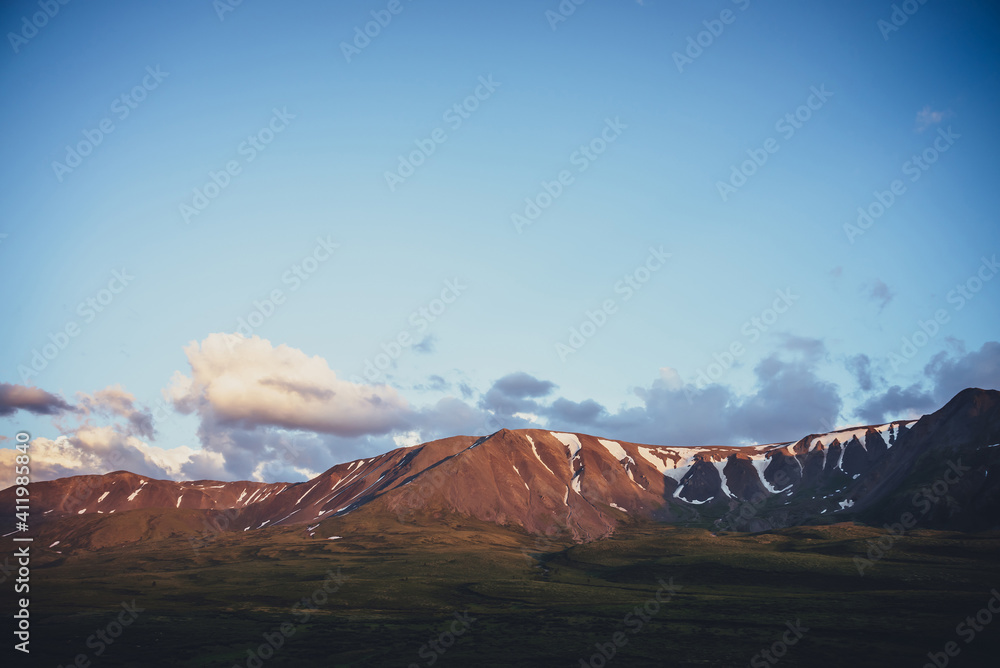 Fototapeta premium Atmospheric mountain scenery with yellow clouds in blue sky. Scenic landscape with illuminating sunset clouds above mountains with snow. Beautiful sunrise in mountains. Snow on rocks in sunlight.
