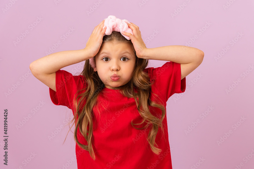A small beautiful girl with curly hair on a purple background holds her head and puffed out her cheeks. A small beautiful girl with curly hair on a purple background holds her head and puffed out her cheeks.