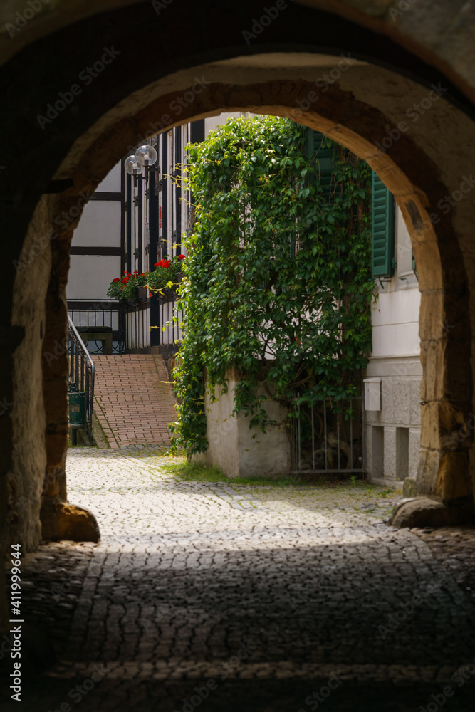 Fototapeta premium dark medieval archway leads into daylight with truss house and cobble stone street of town, Tecklenburg, Germany